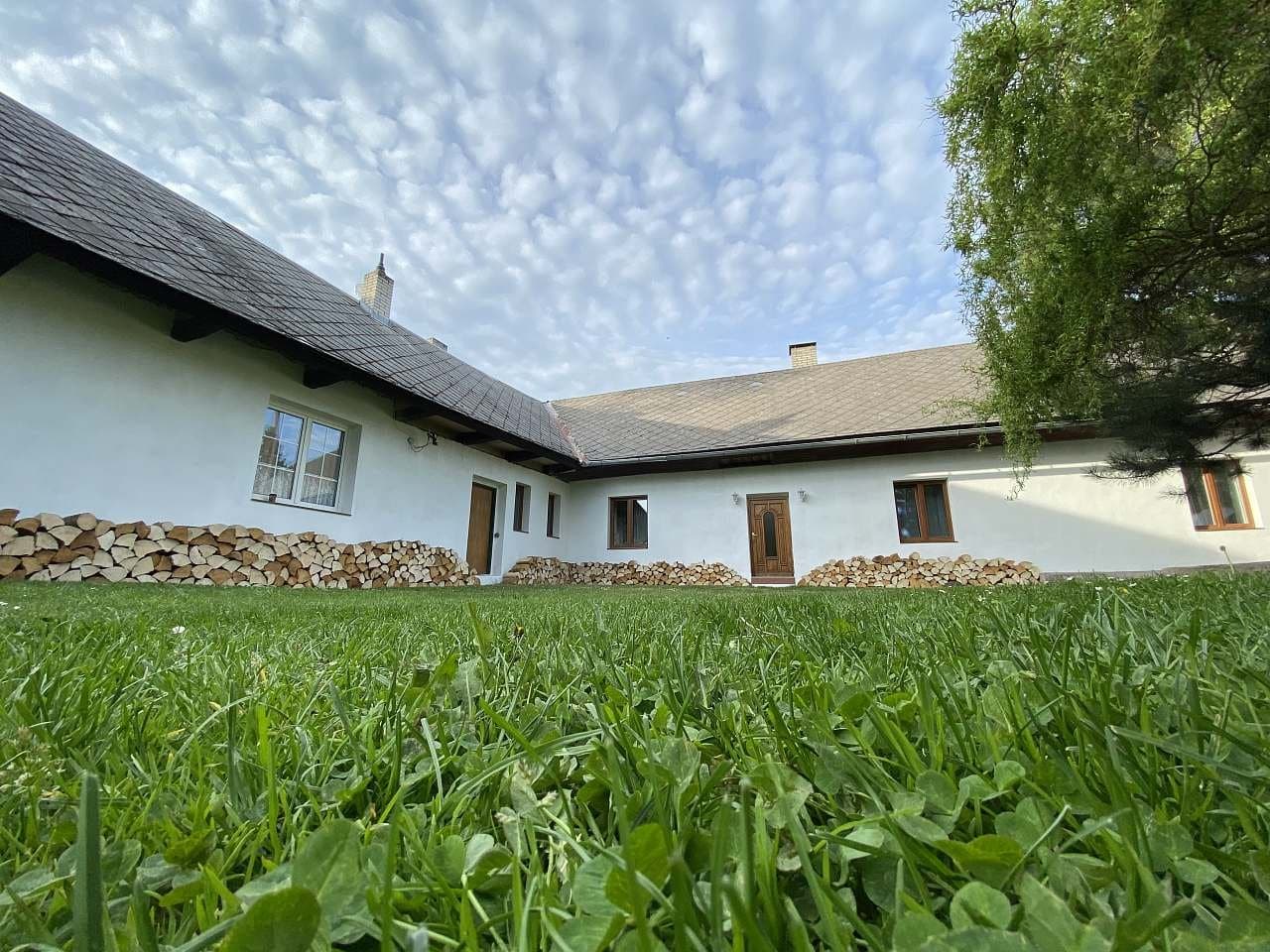 Bílý statek s břidlicovou střechou a naštípaným dřevem u zdi, v popředí tráva